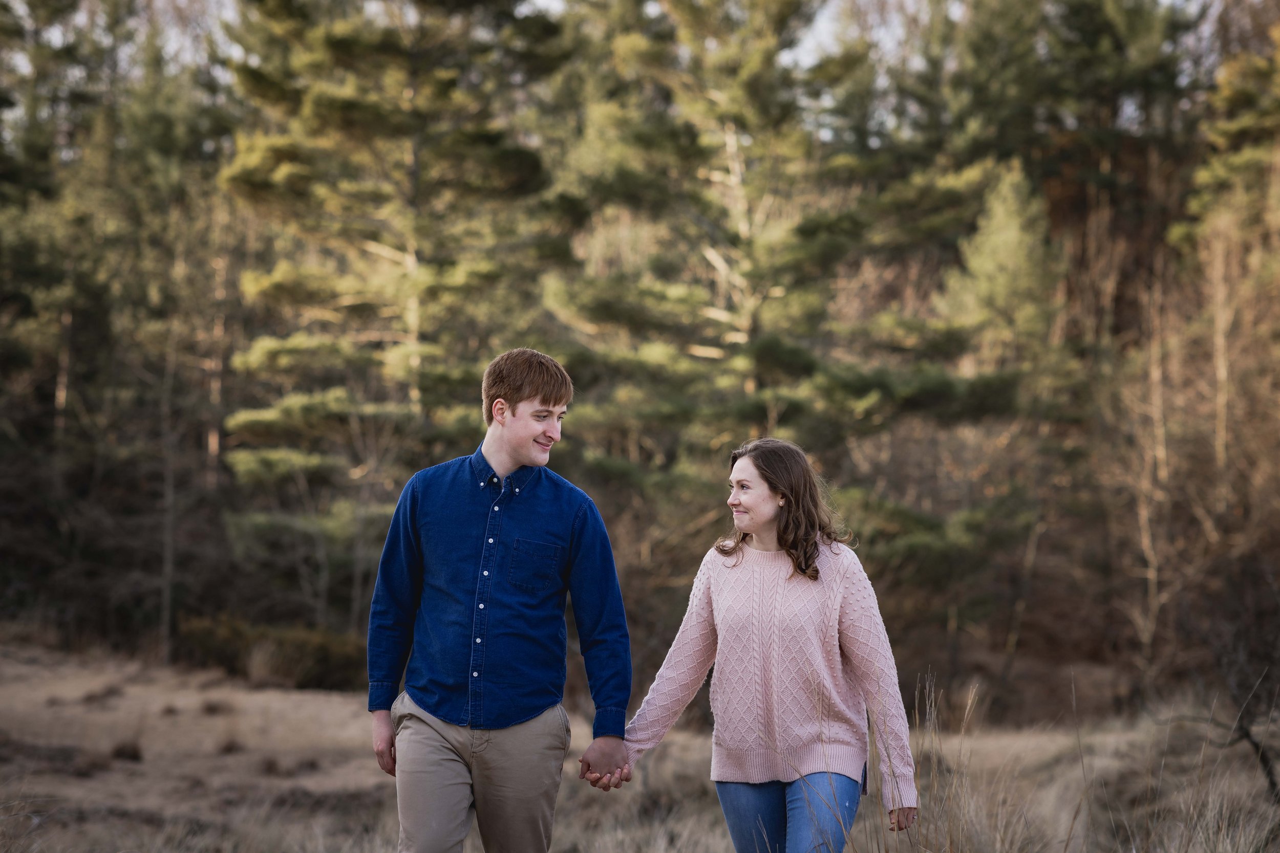 An engaged couple holding hands with tall green pines in the background 