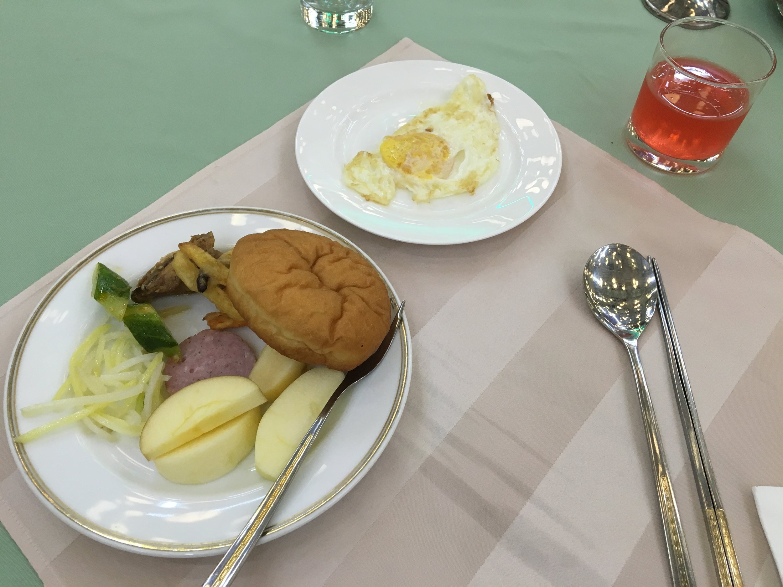 Breakfast of various pickles, spam, apple slices, and fried egg with a cup of artificial red apple juice.