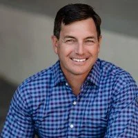 A smiling man with dark hair wearing a blue checkered shirt, posing against a neutral background.