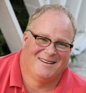 A smiling man with glasses and short light hair, wearing a red polo shirt, outdoors.
