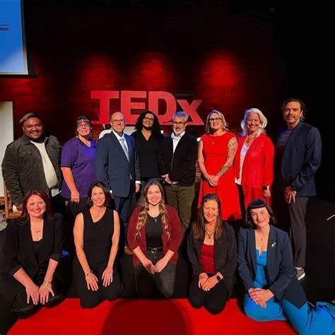 Group of ten diverse individuals standing and kneeling on stage with a red TEDx logo background, smiling at the camera.
