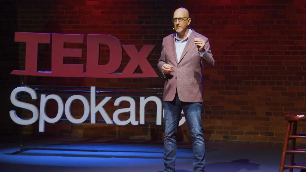 Man giving a presentation on a stage with a brick wall background, standing next to large red TEDx Spokane sign.