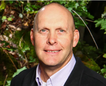 A man with a bald head and blue eyes smiling, wearing a dress shirt and blazer, standing outdoors in front of greenery.