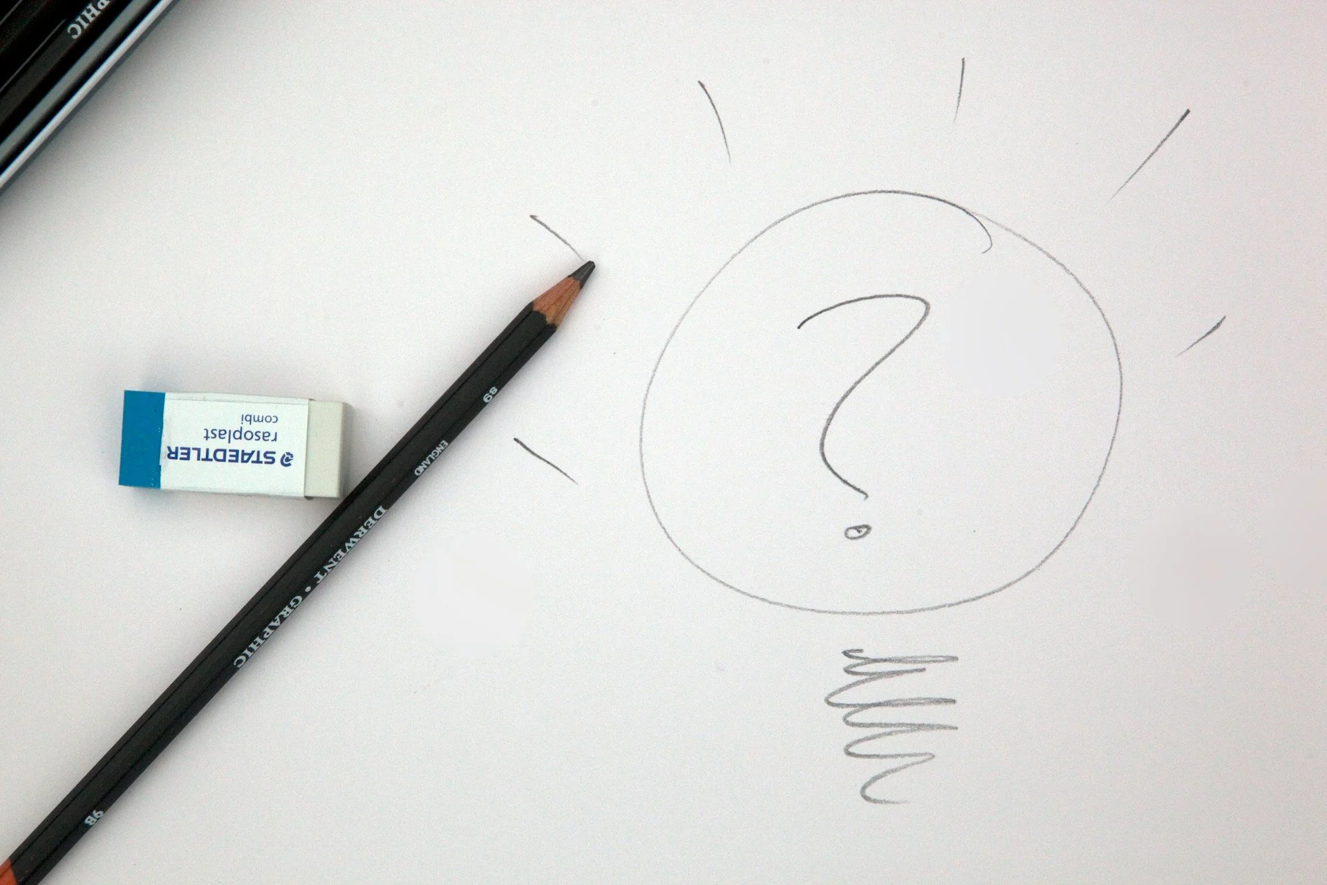 A photo of a white board with a pencil and rubber laying on it. Drawn on the paper is a question mark within a bulb.