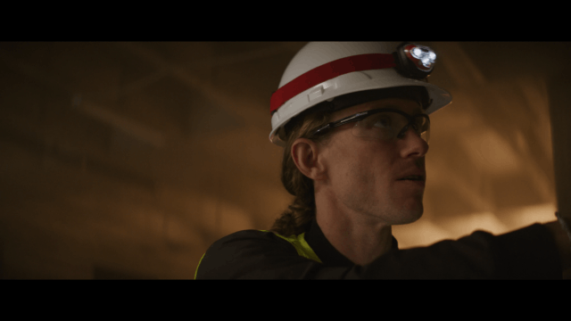 Focused construction worker wearing a hard hat with headlamp, safety glasses, and high-visibility vest, inspecting a structure in a warmly lit industrial environment, symbolizing safety, precision, and field expertise.