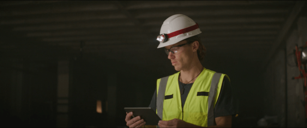 Construction worker wearing a safety helmet, headlamp, and high-visibility vest using a tablet in a dimly lit industrial environment, highlighting the use of digital tools in modern site inspections and project management.