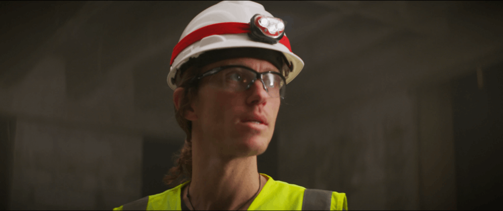Close-up of a construction worker wearing a safety helmet with a headlamp, high-visibility vest, and protective glasses, standing in a dimly lit industrial or construction site, representing safety and focus on the job.