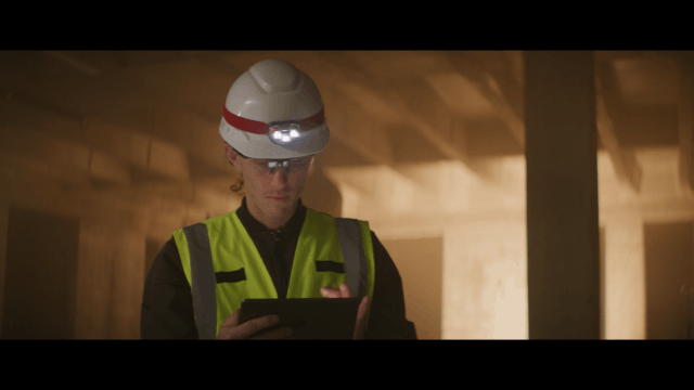 Construction worker in a high-visibility vest and hard hat with headlamp using a tablet in a warmly lit industrial setting, representing modern site management and digital workflow solutions.