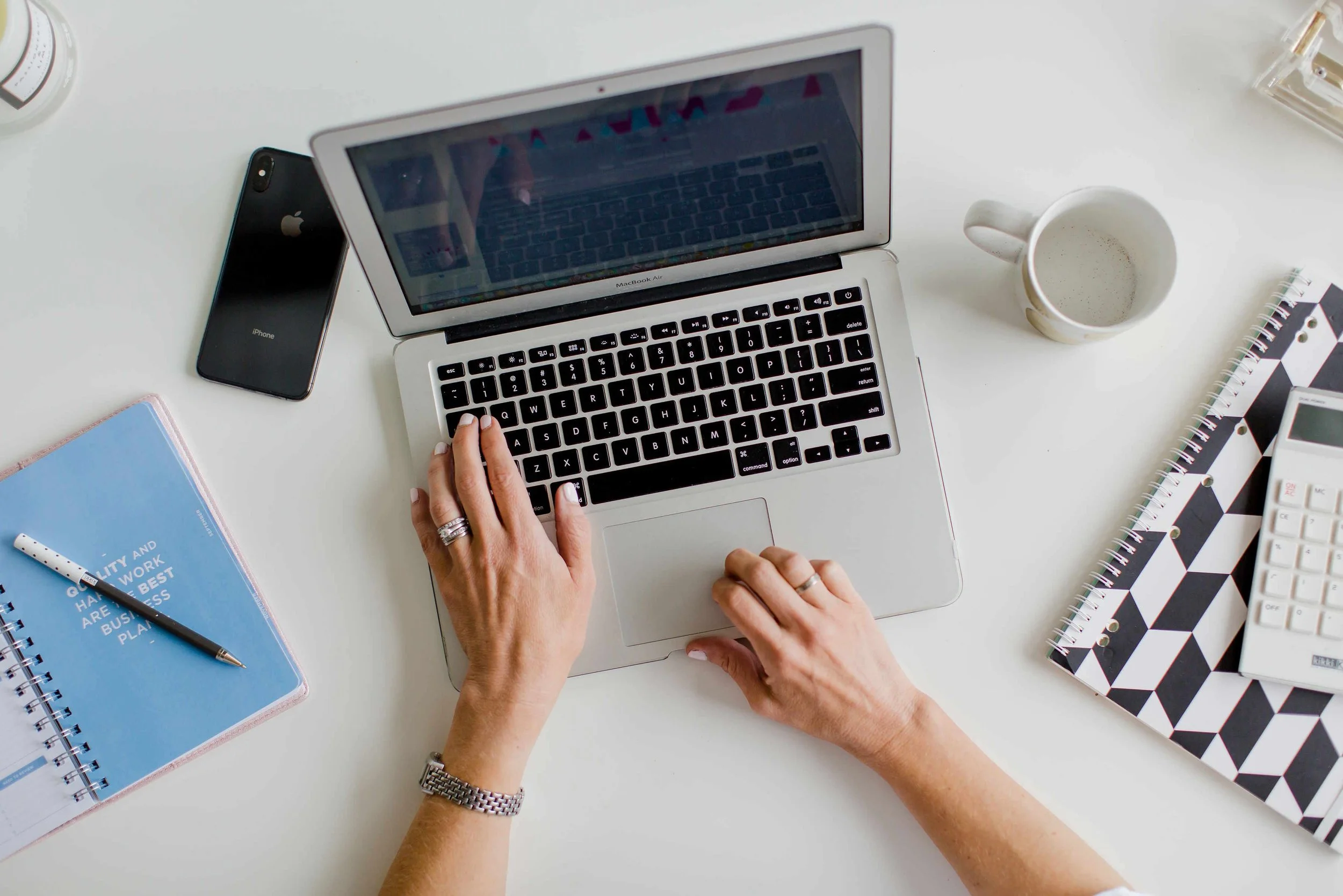 Top-down view of a workspace with a person's hands typing on a silver MacBook Air laptop, surrounded by a black iPhone, a blue notebook with a pen, a white mug with coffee, and various notebooks and office supplies on a white desk.