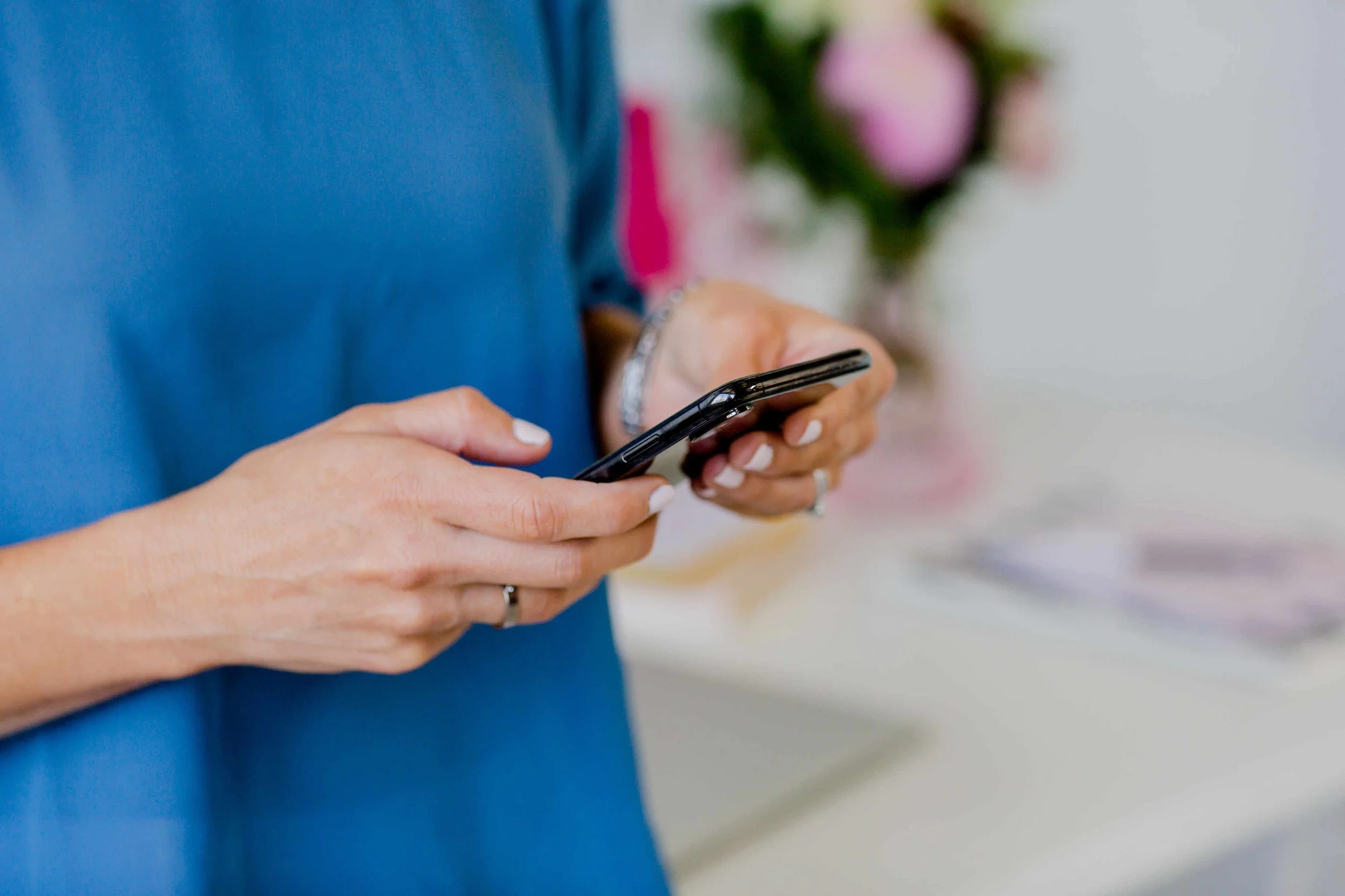 Close-up of a person wearing a blue shirt and holding a smartphone with both hands, in front of a blurred background with pink and green flowers.