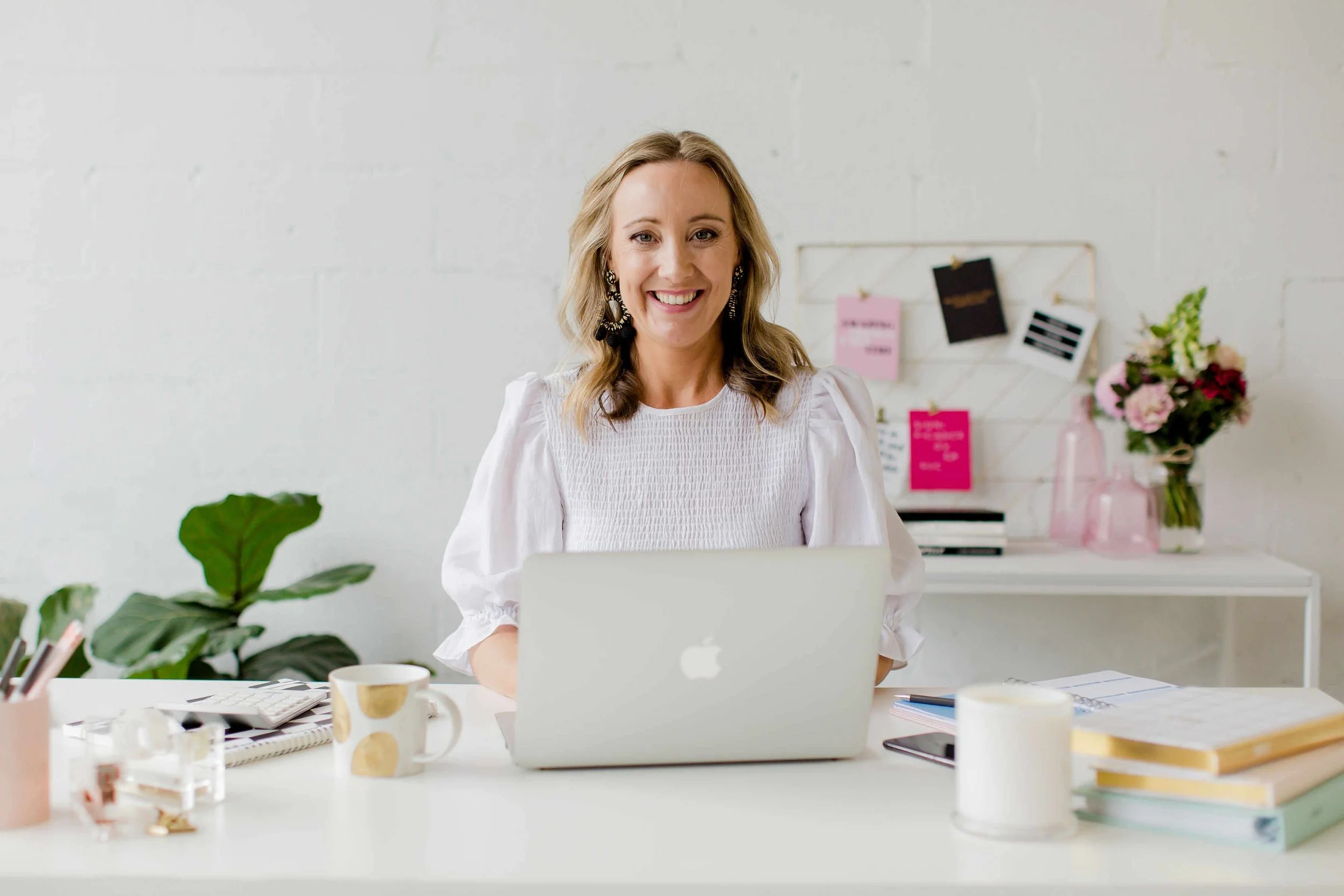 A woman with blond hair, wearing earrings and a white blouse, sitting at a desk with a laptop, colorful sticky notes on the wall behind her, and various office supplies including a notebook, cups, and a plant in the foreground.