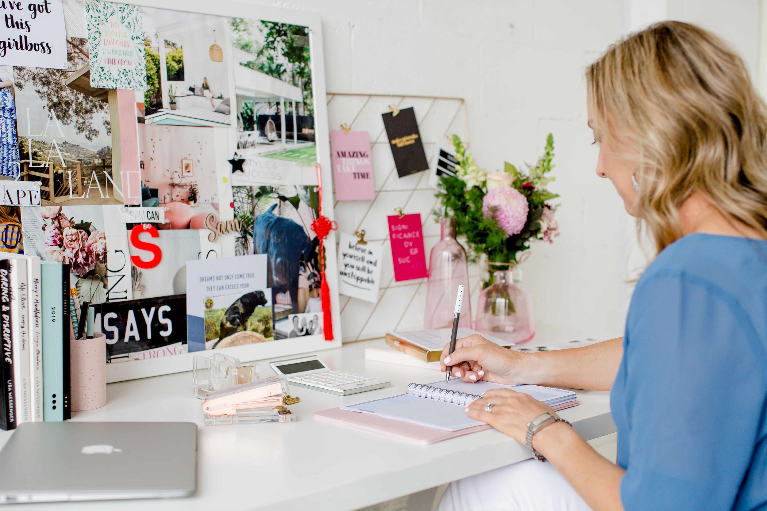 A woman sitting at a white desk, writing in a planner, with a large decorated bulletin board behind her filled with photos, quotes, and decor, and a vase of flowers on the desk.