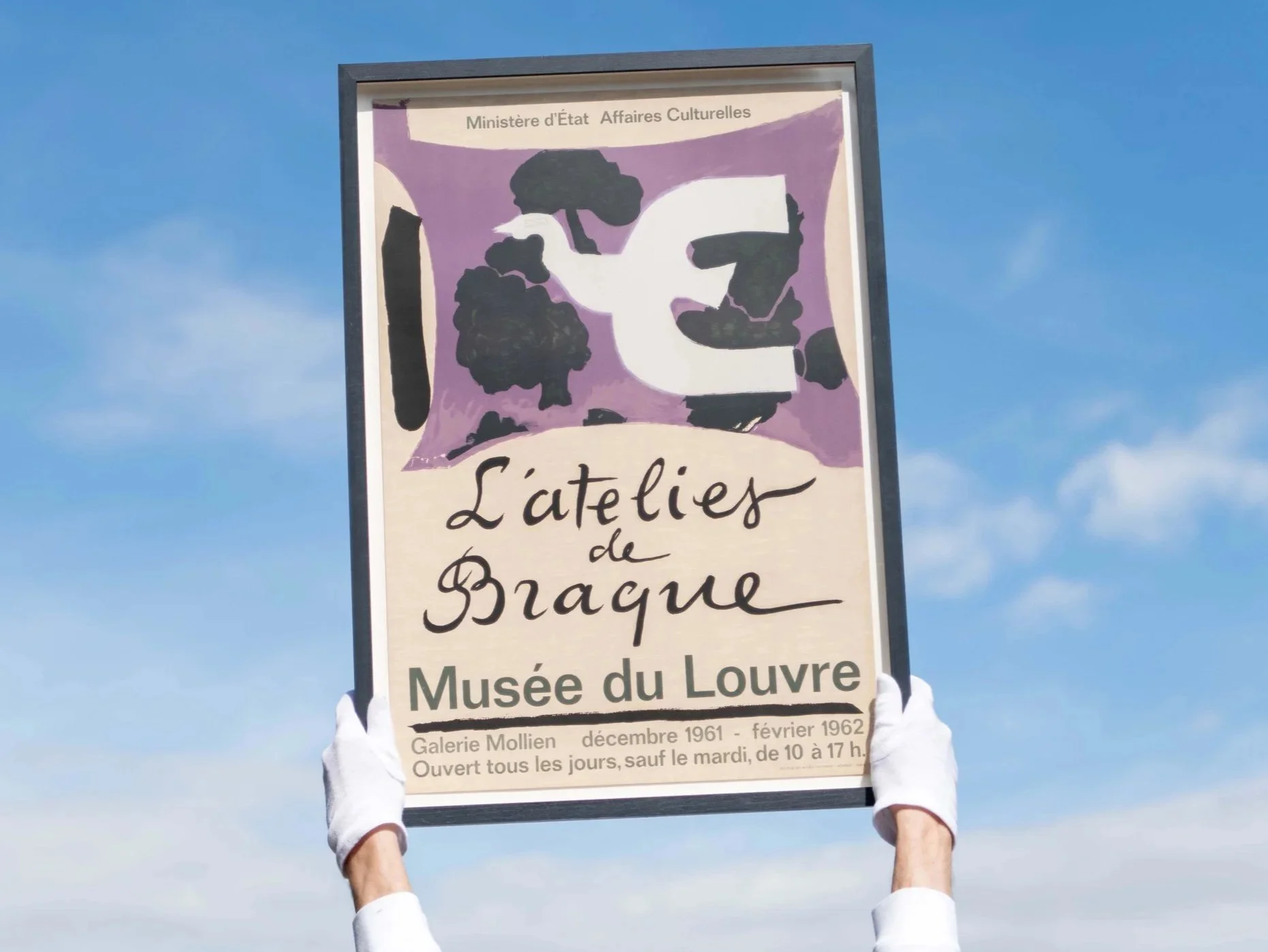 A person wearing white gloves holds a sign above their head against a blue sky background. The sign is from the Musée du Louvre and features a purple and black abstract design with text in French.