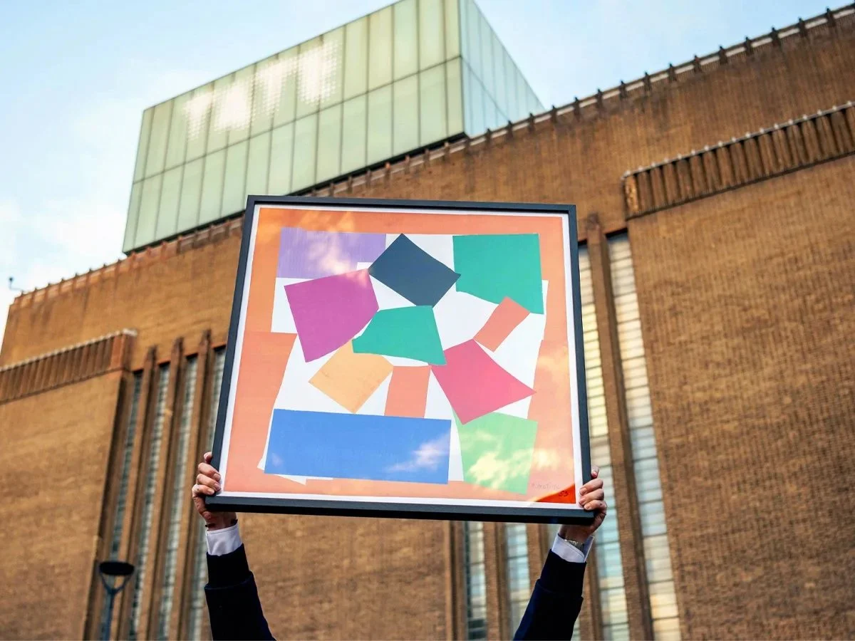 Person holding a colorful abstract artwork in front of a modern brick building with glass windows.