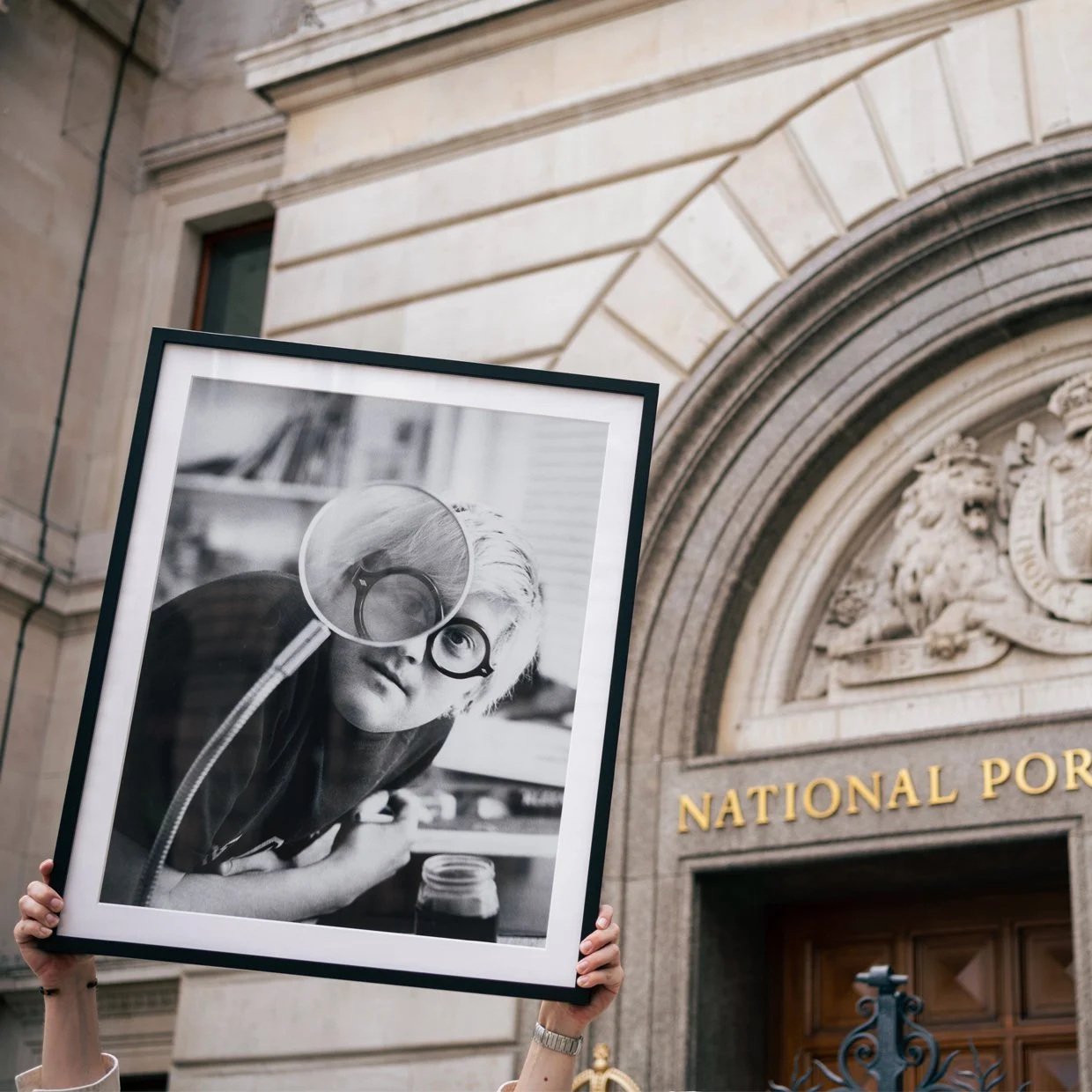 Person holding a framed black and white photograph of a woman with glasses and a magnifying glass, in front of the National Portrait Gallery building.