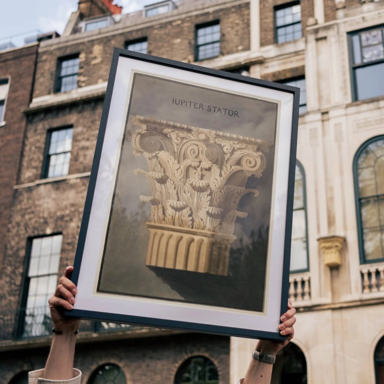 Person holding an illustration of an Ionic column capital labeled "JUPITER-STATOR" outdoors, with a brick building in the background.