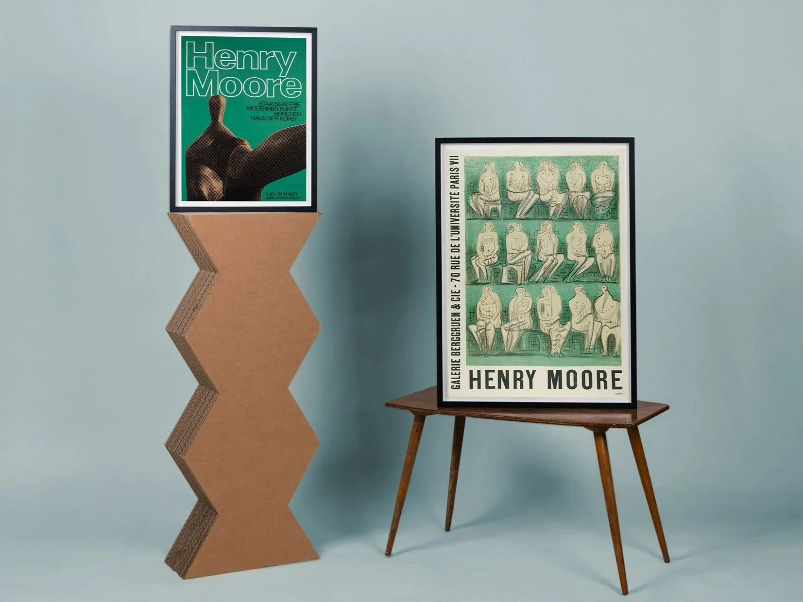 Two framed art posters by artist Henry Moore. The left poster features a large green background with a black sculpture and text. The right poster shows a green illustration of multiple seated human figures in black outline with text along the left si