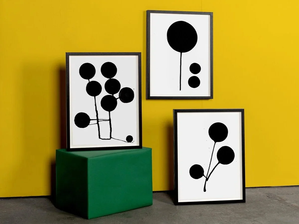 Three black and white abstract balloon artworks in black frames against a yellow wall, with one artwork resting on a green block.