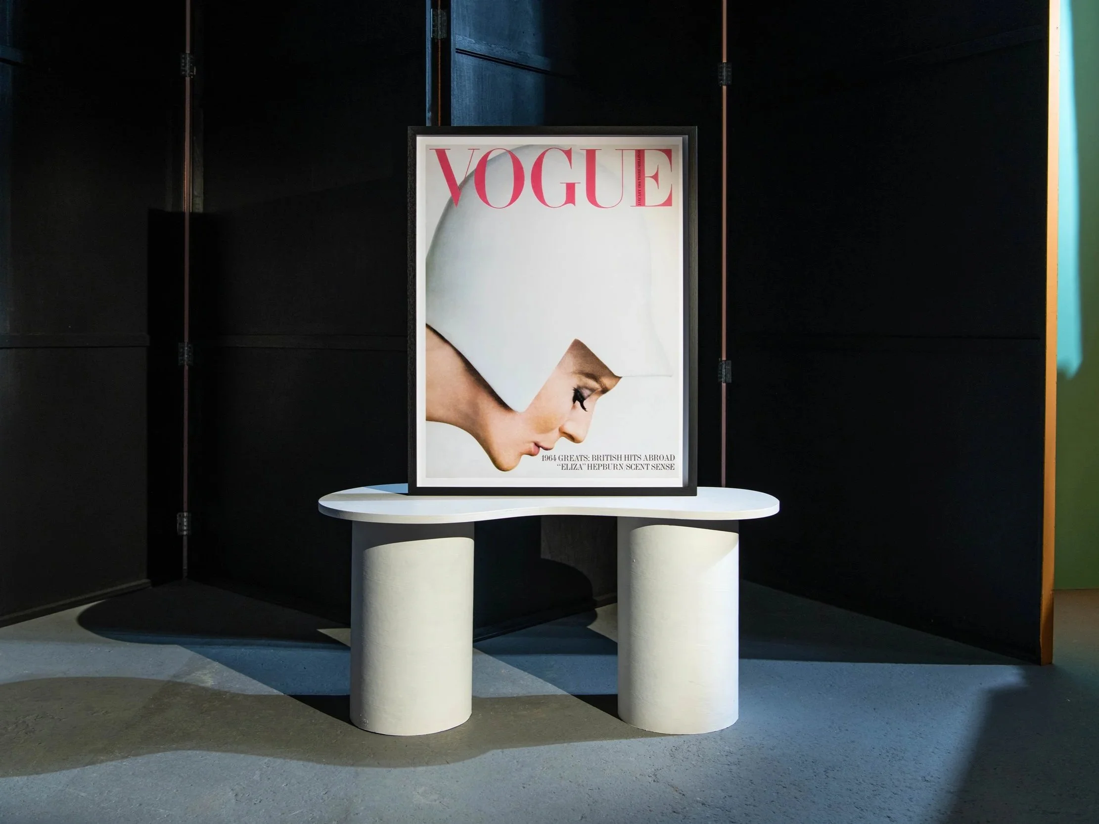 Framed magazine cover of Vogue featuring a woman with white geometric head covering and makeup, displayed on a white pedestal against dark background.