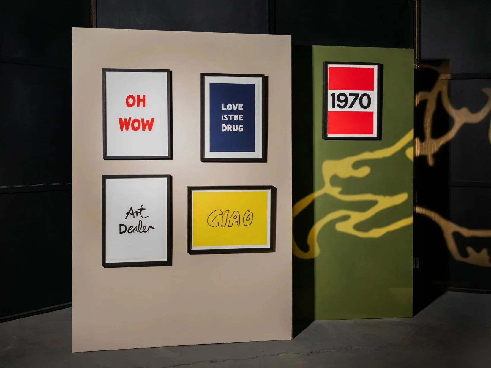 Gallery wall with five framed graphic posters. Top left: red text 'OH WOW,' top middle: blue background with white text 'LOVE IS THE DRUG,' bottom left: white background with black handwritten 'Art Dealer,' bottom right: yellow background with black 