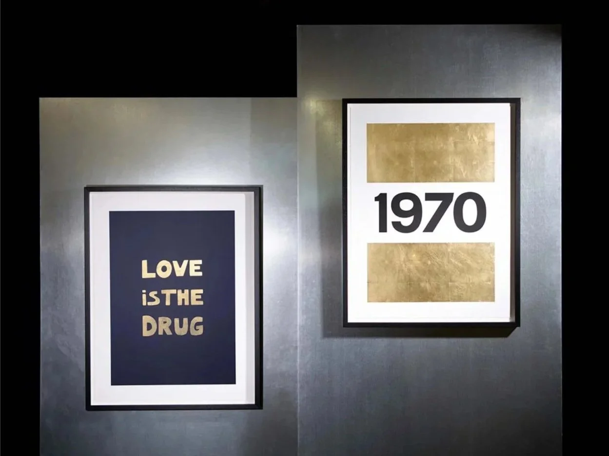 Two framed posters on a wall, one with the words 'Love is the drug' in gold letters on a dark background and the other with '1970' in bold black text, surrounded by gold rectangles.