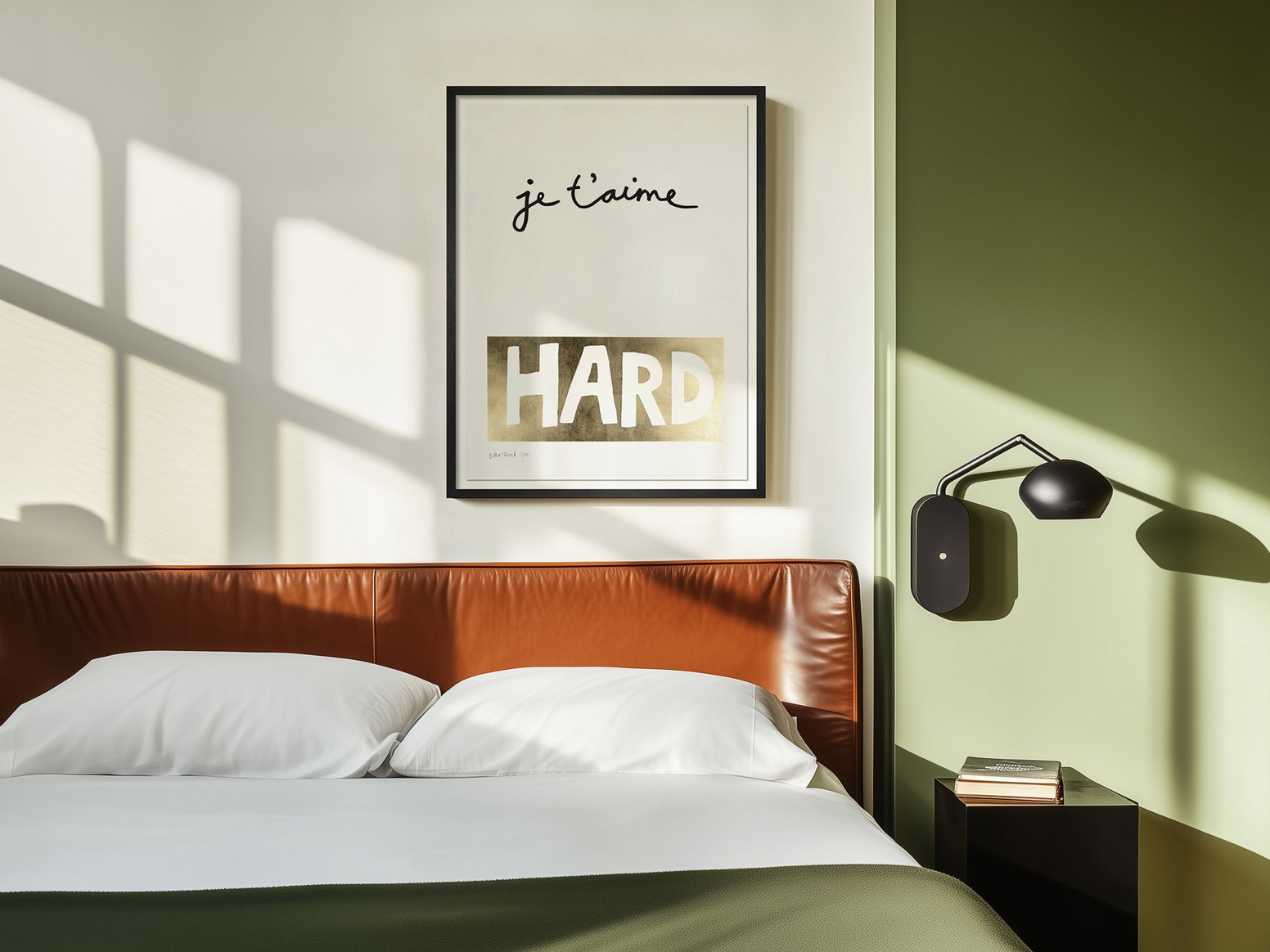 A bedroom with a brown leather headboard, white pillows, a black bedside table with books, a wall-mounted black lamp, a framed art print that says 'je t'aime' and 'HARD', and a green accent wall.