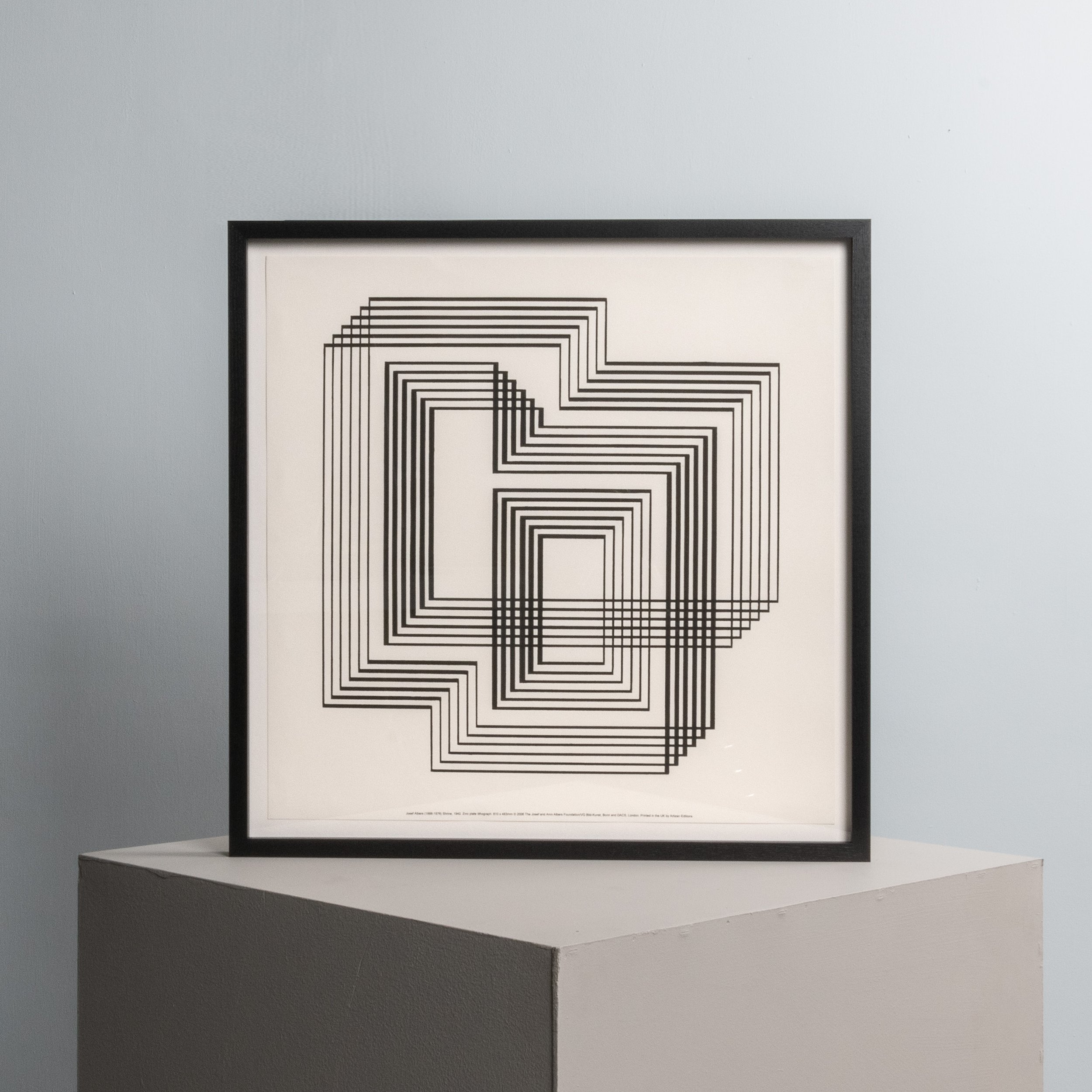 A framed artwork with black geometric lines forming nested squares and rectangles on a white background, displayed on a white pedestal against a light grey wall.