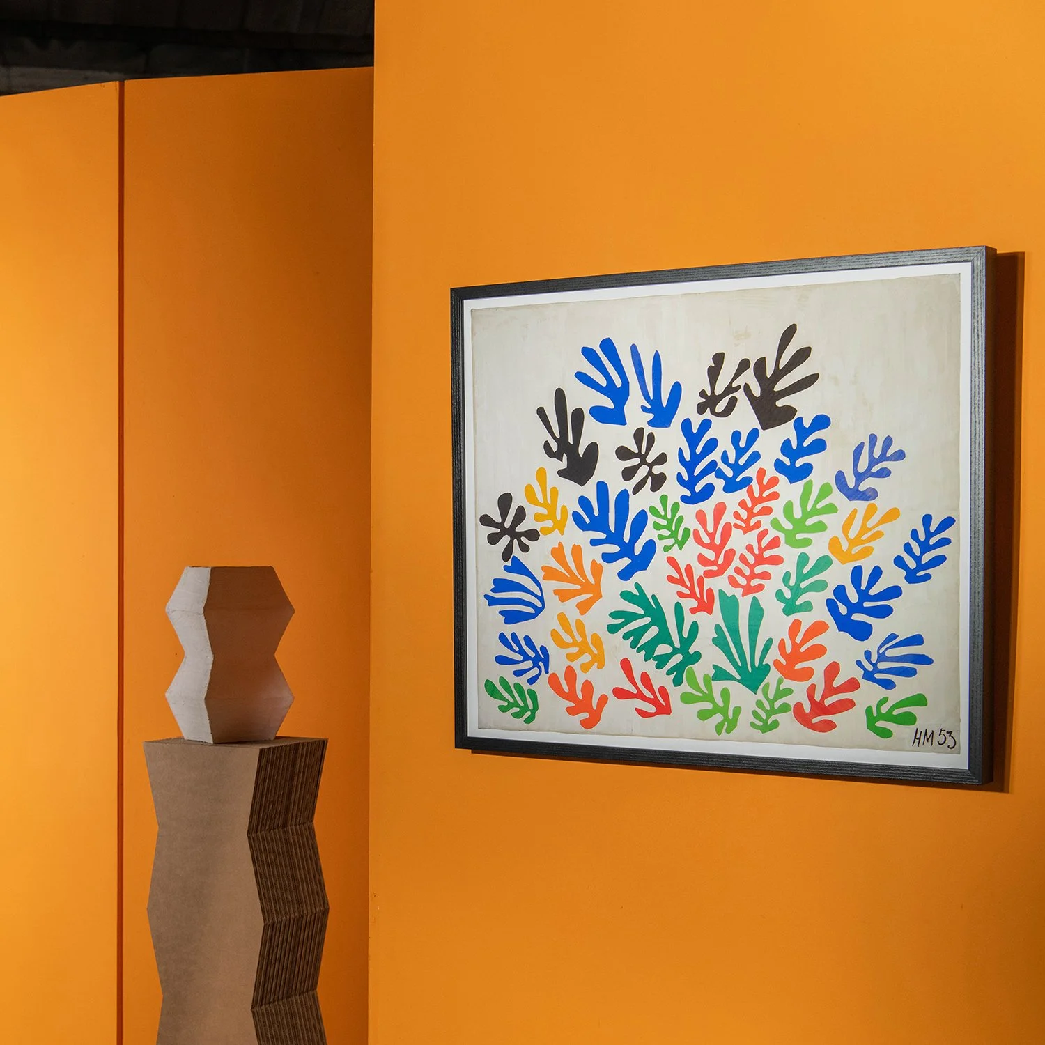 Colorful abstract art featuring numerous hand-shaped designs in blue, green, red, yellow, and black, arranged on a white background, framed and displayed on an orange wall.