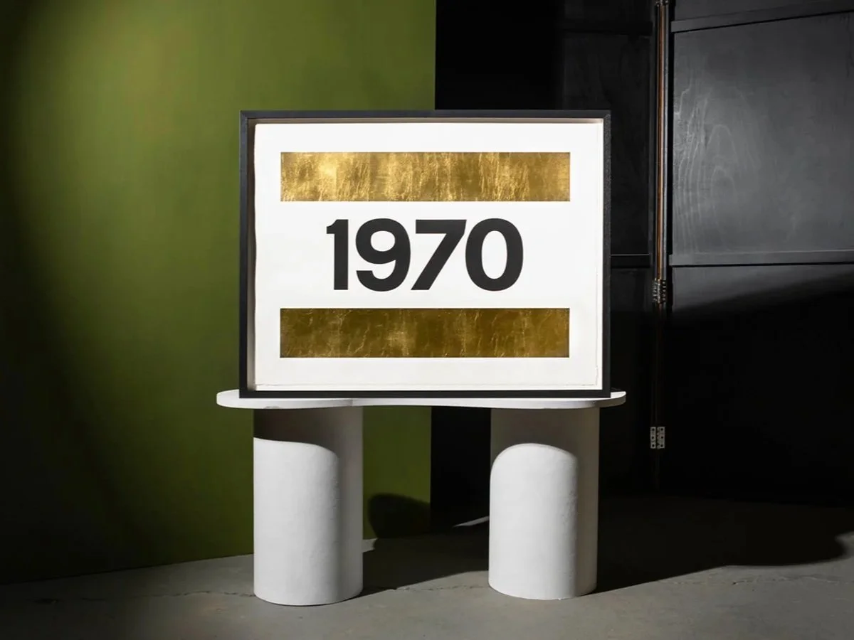 A display sign with the year 1970, decorated with gold accents, mounted on a minimalist white stand.