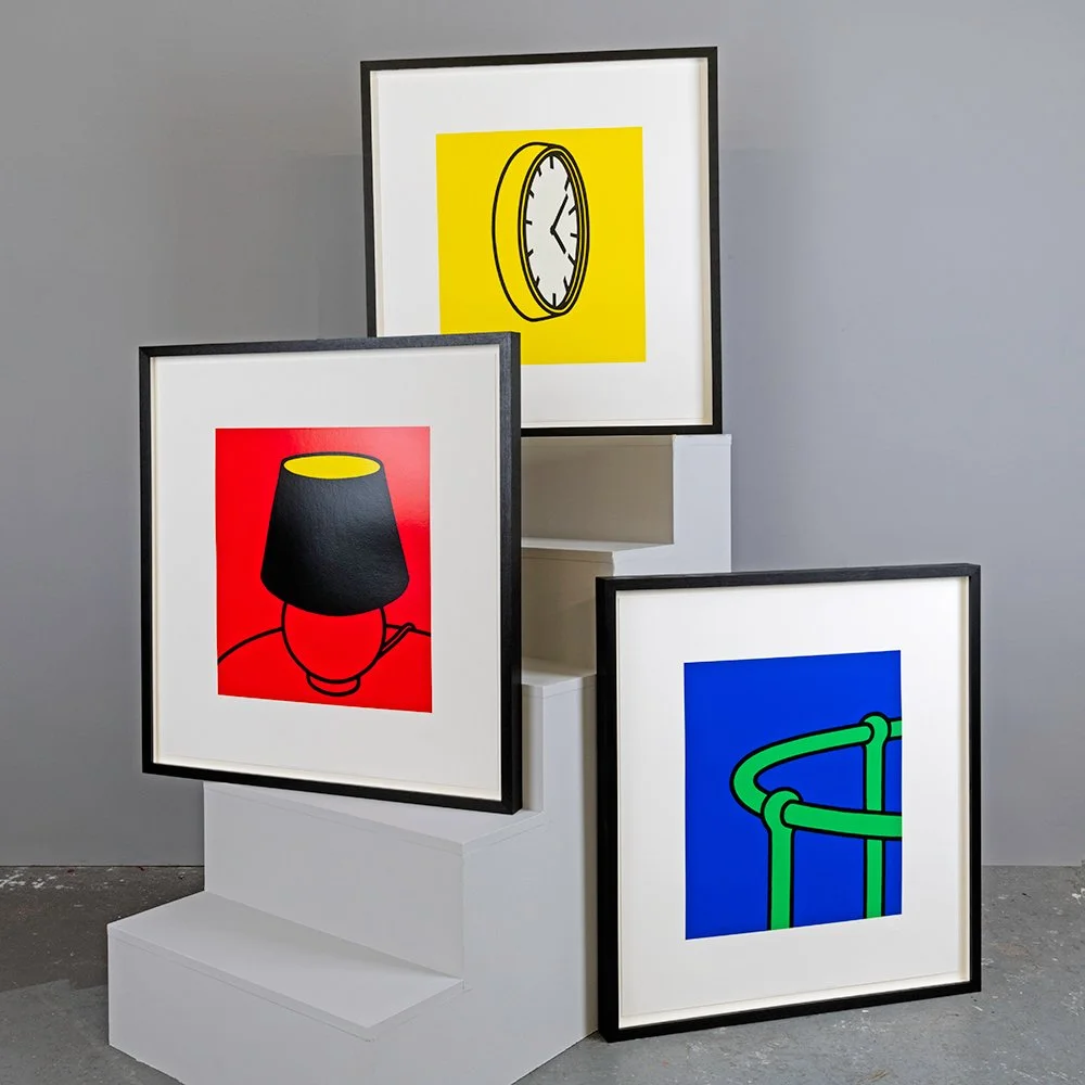 Three framed pop art images on white display stands against a gray wall. The images depict a clock, a lamp, and abstract green tube shapes on colorful backgrounds.