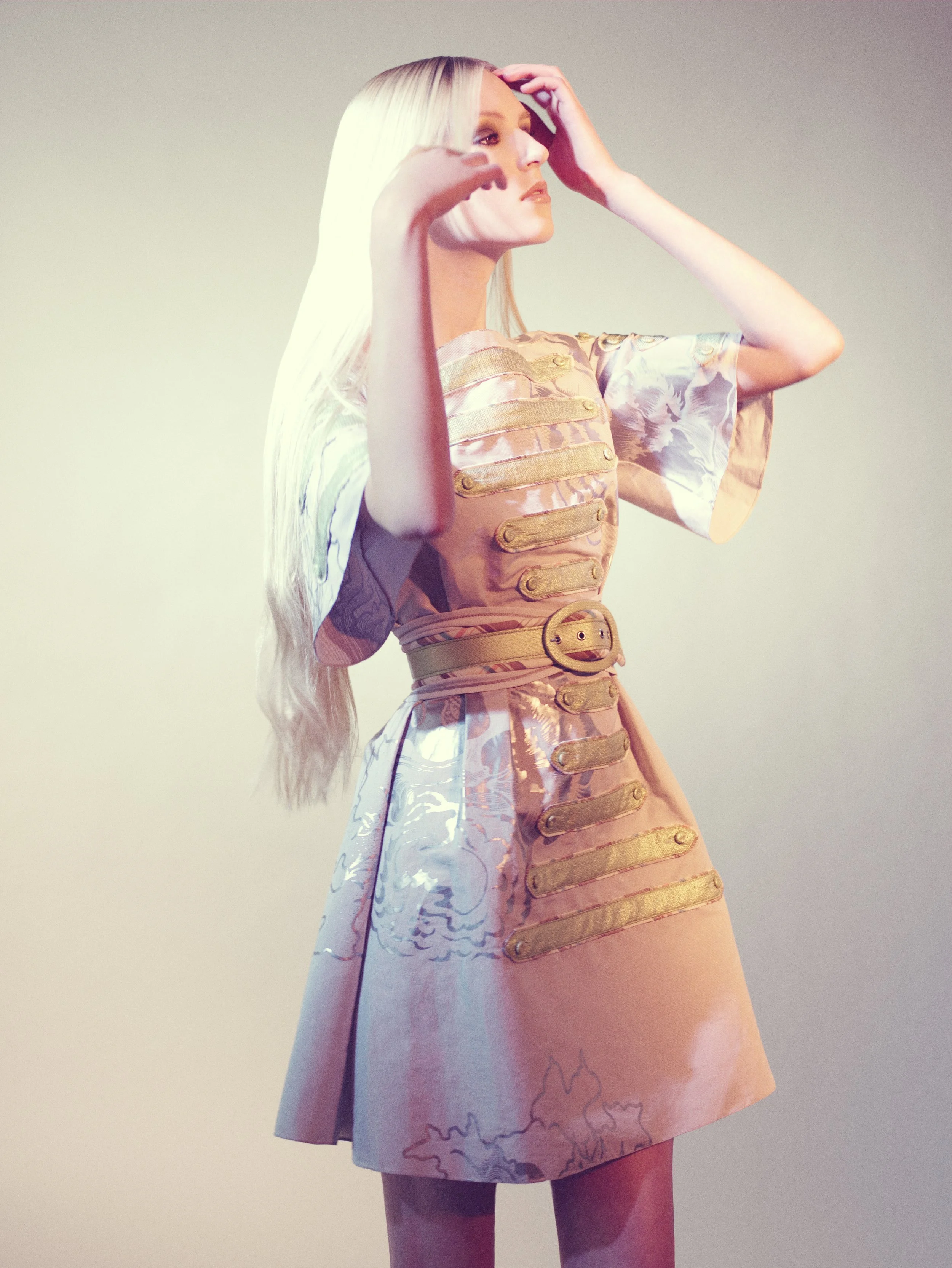 Woman with long, platinum blonde hair wearing a pastel-colored dress with gold details, standing against a plain light-colored background.