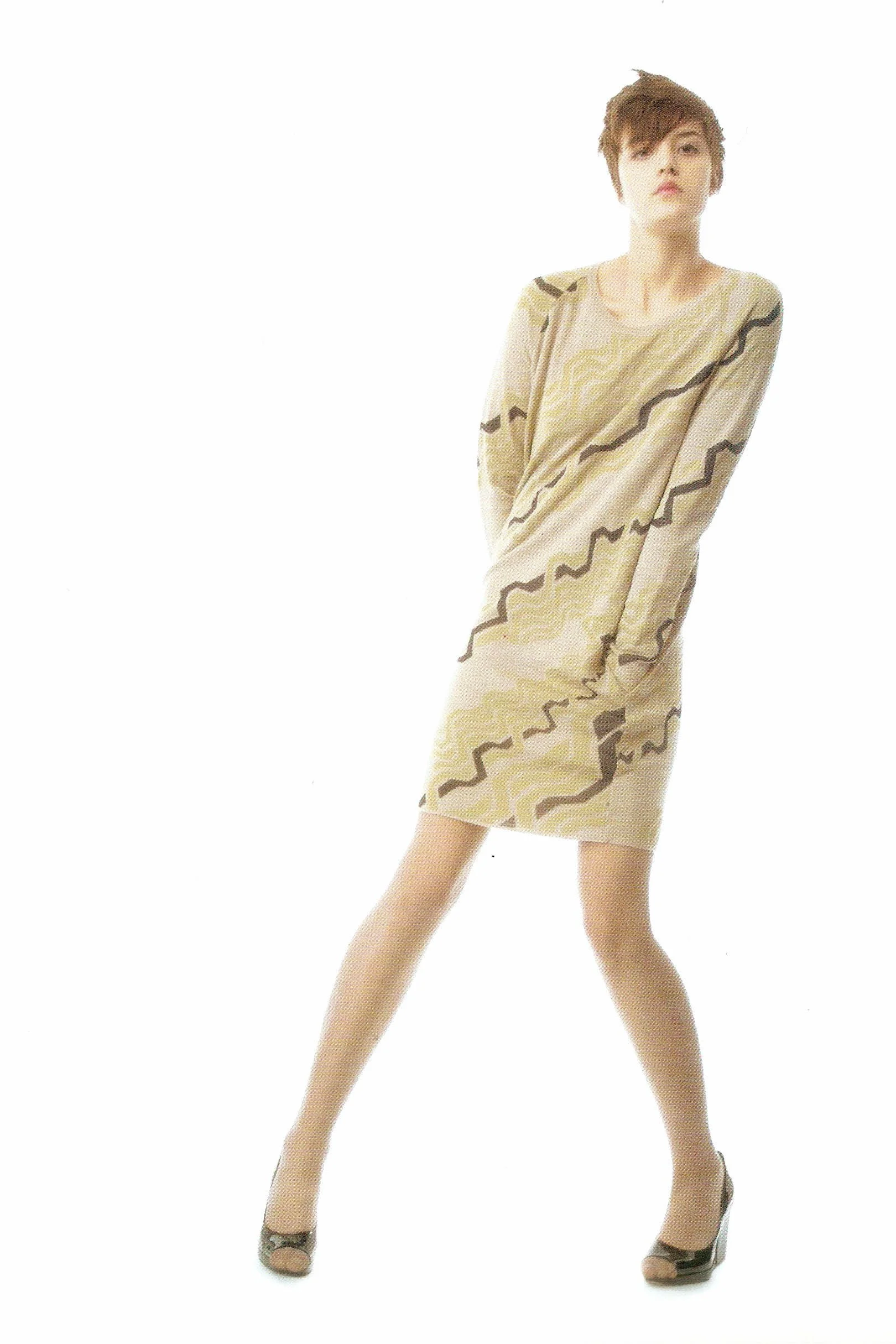 A woman in a long beige dress with dark wavy line patterns, black heels, standing with legs apart and arms crossed, looking at the camera against a white background.