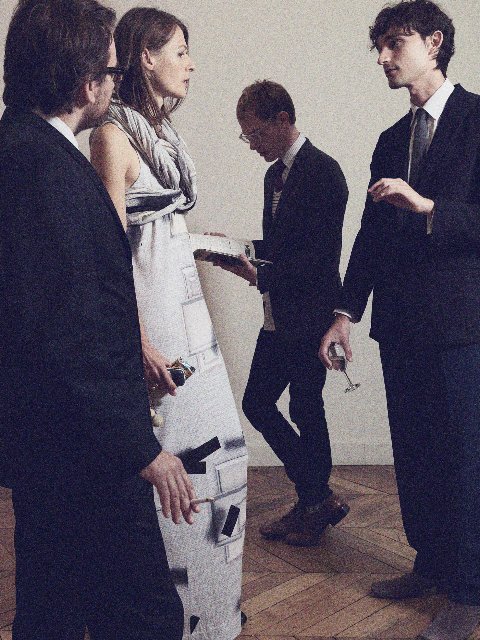A group of five young adults dressed in formal attire, engaged in conversation in an indoor setting with wooden flooring.