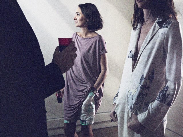 Two women standing and talking with a person holding a red cup. One woman is wearing a loose purple dress, and the other is in a white shirt with a print.