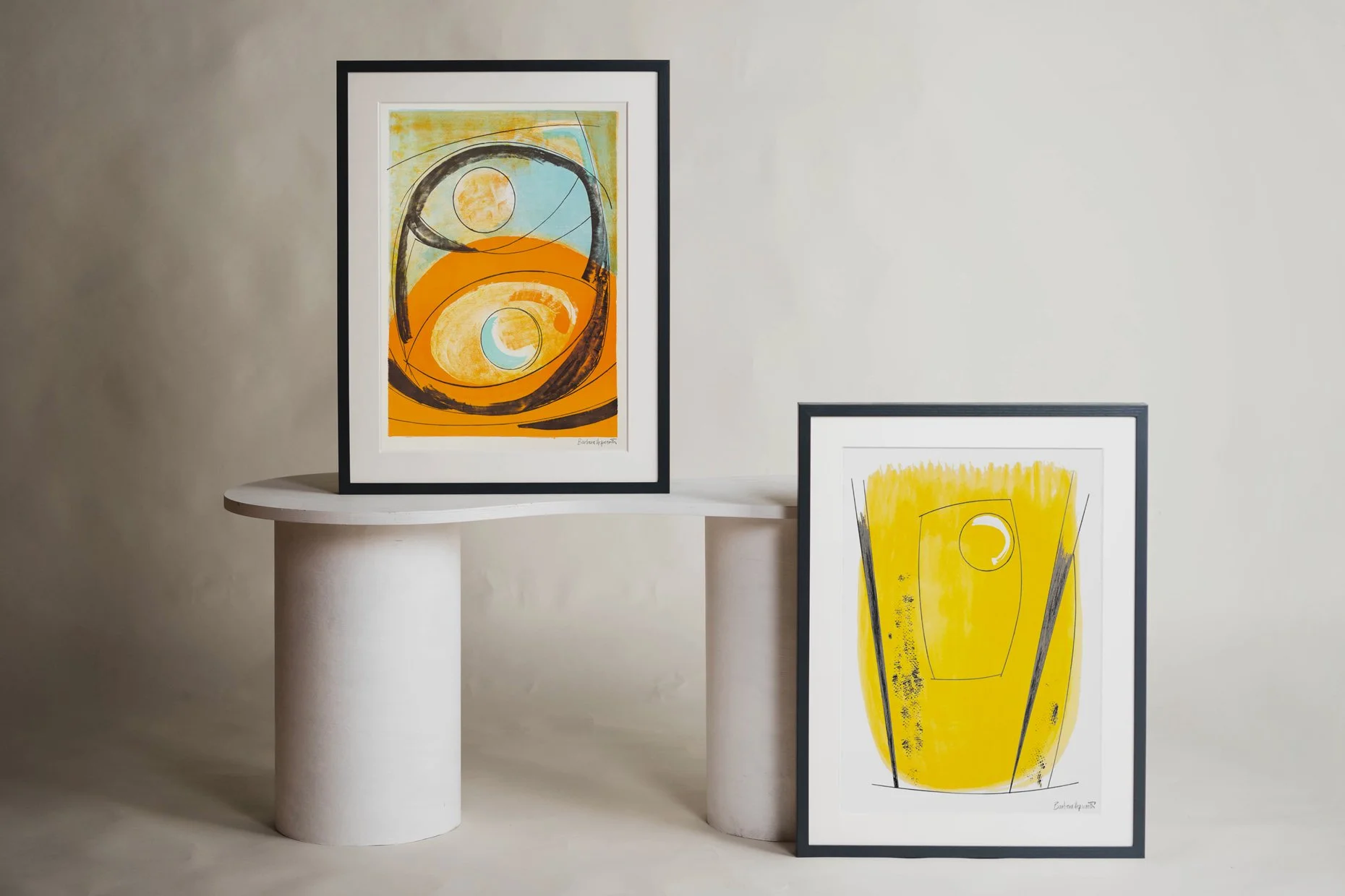 Two abstract art prints with yellow and orange tones, framed in black, displayed on white pedestals and against a light-colored wall.