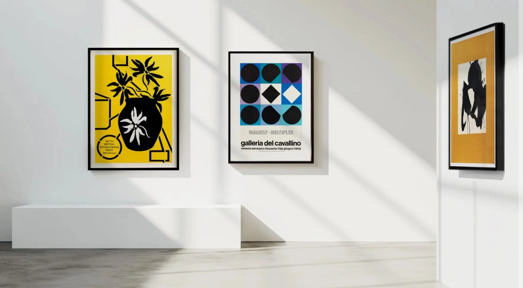 Gallery wall with three framed art posters in a bright, white room. The left poster features a black and yellow design with a plant in a vase. The middle poster has a pattern of black circles, a diamond, and text that reads "VASARELY - MULTIPLES gall