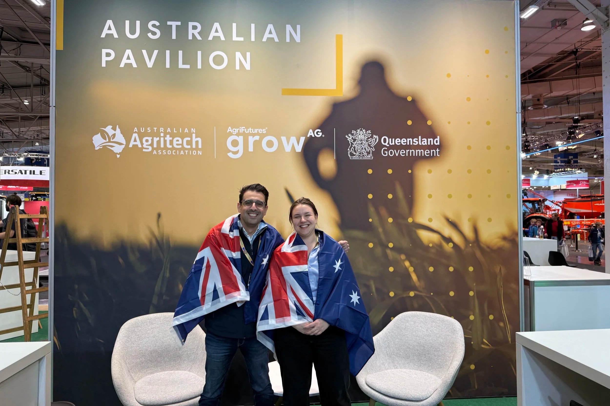 Two people smiling at the Australian Pavilion, wearing Australian flag capes, standing in front of a backdrop with the text 'Australian Pavilion' and logos of Australian agricultural organizations and the Queensland government, at an indoor exhibition or trade show.