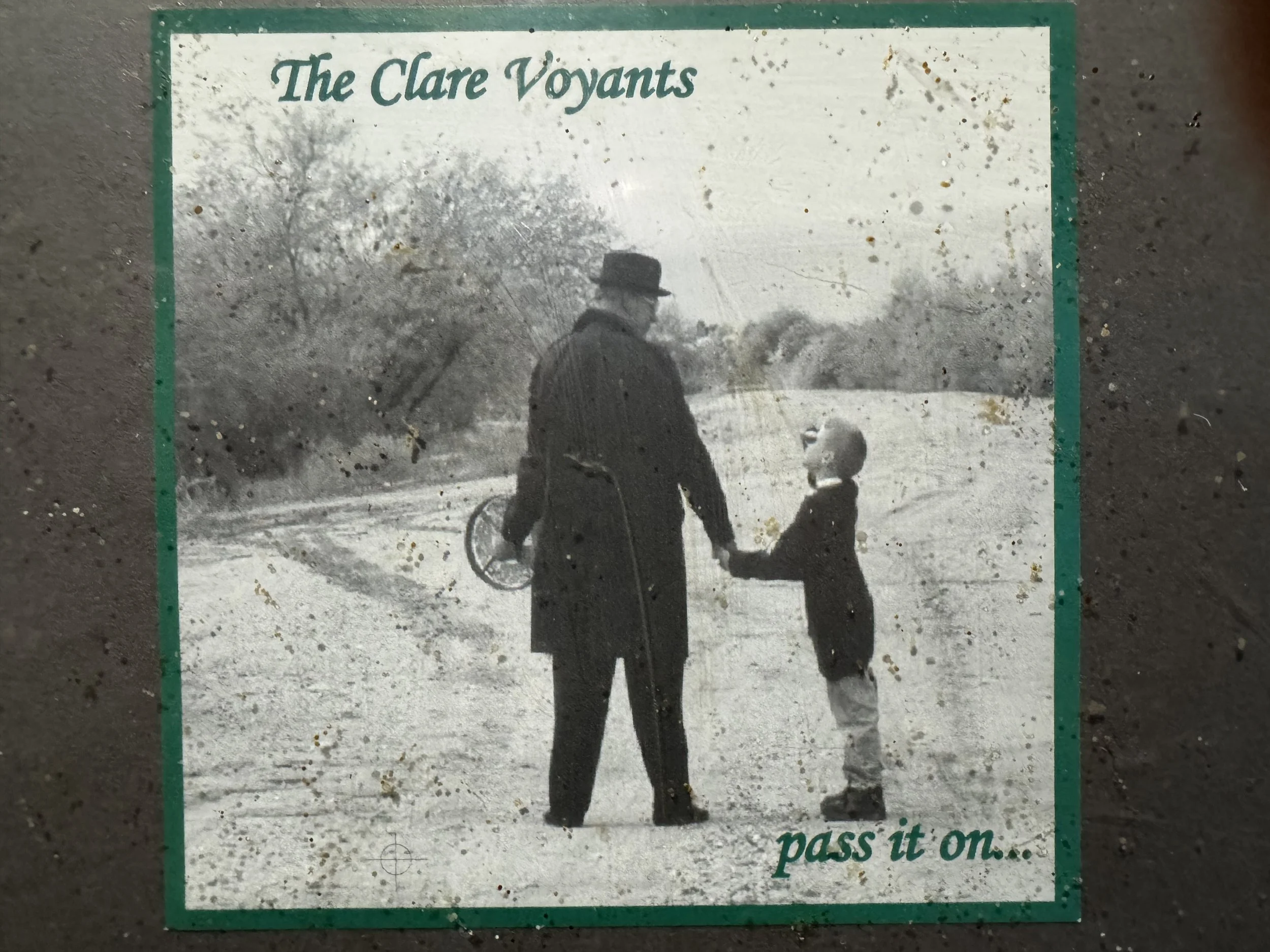 Black and white photograph of a man in a hat and coat holding hands with a child, walking outdoors with trees and an open field in the background. Text reads 'The Clare Voyants' and 'pass it on...'