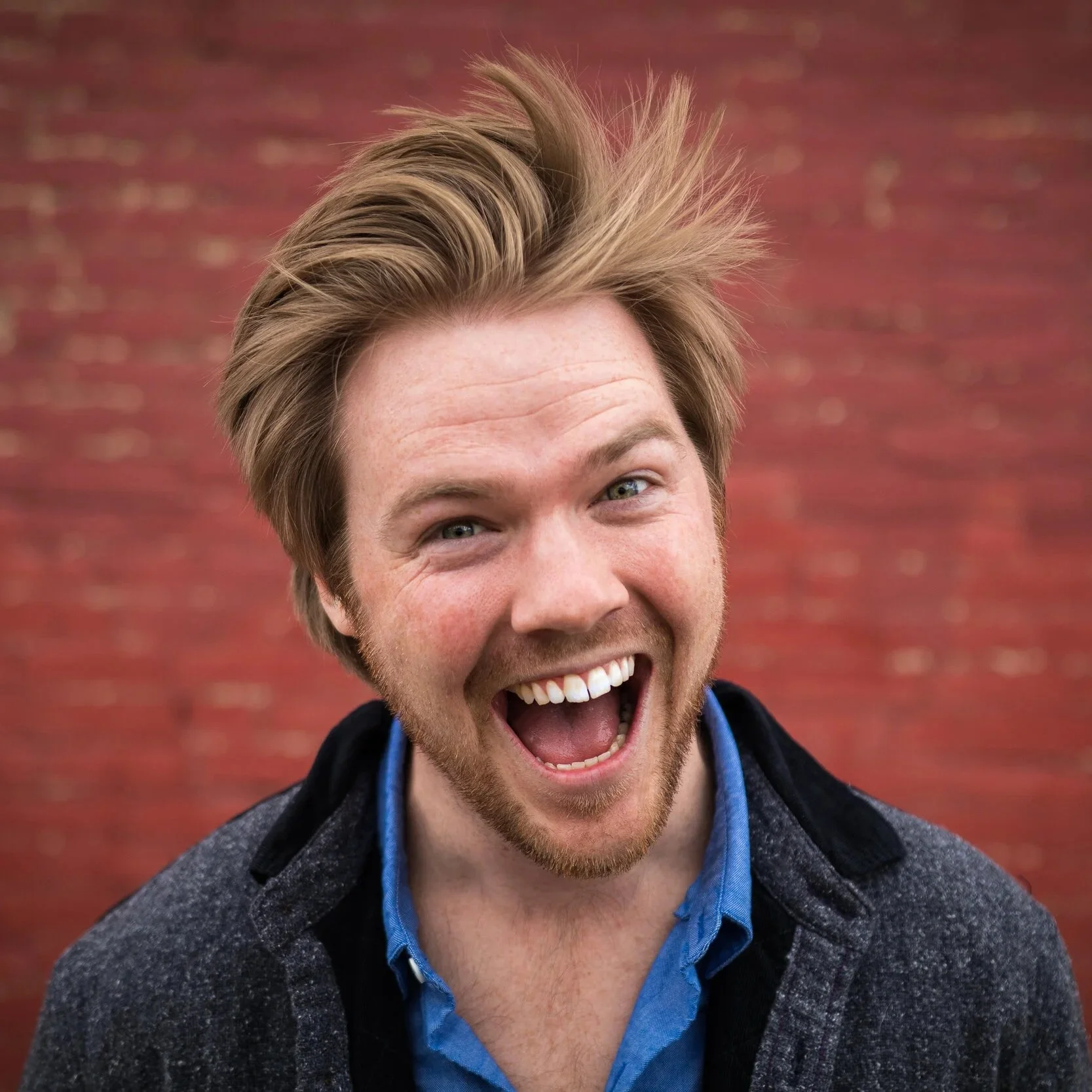 A young man with messy blonde hair, a beard, and wearing a blue shirt and a dark jacket, smiling widely outdoors in front of a red brick wall.