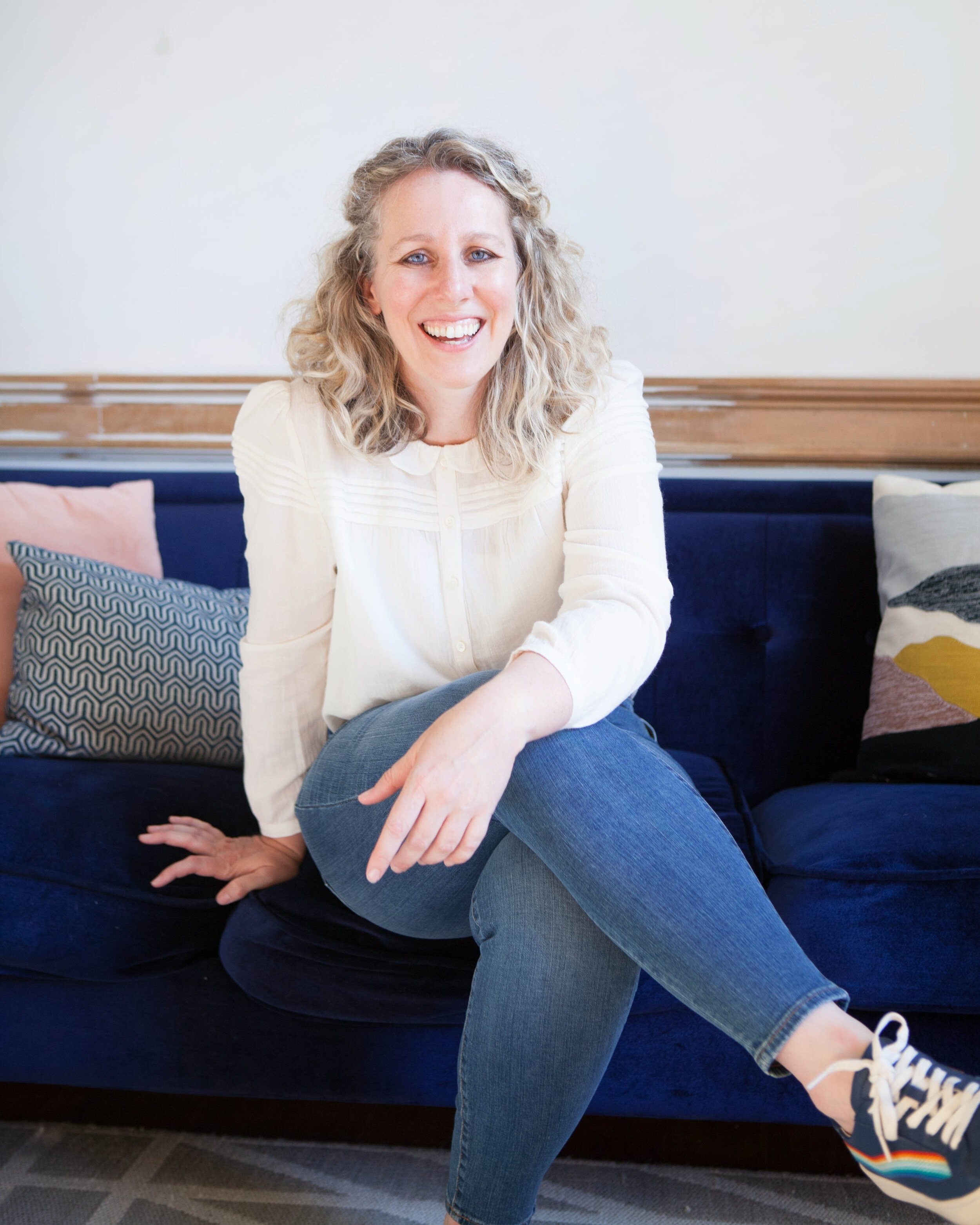 A smiling woman, Kate Hagborg, LMHC, sitting on a couch and offering therapy for people dealing with imposter sydrome.