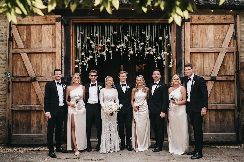 Line of bride, groom and bridesmaids standing in front of a flower arch at Tythe by The Flower Story