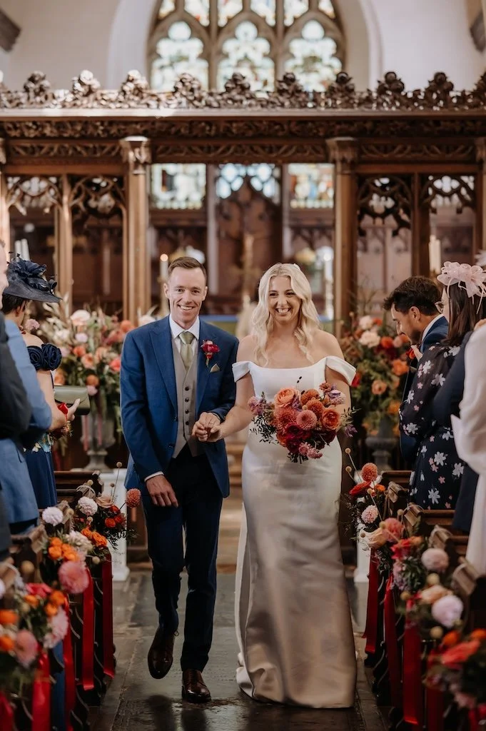 Just married couple walking down the aisle at St Marys church surrounded by colourful wedding flowers
