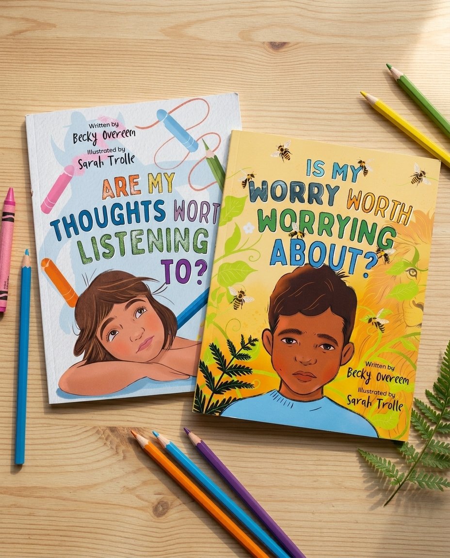 Two children's books about worries and thoughts, one with a girl resting her head and the other with a boy, surrounded by crayons, pens, and green fern leaves on a wooden table.