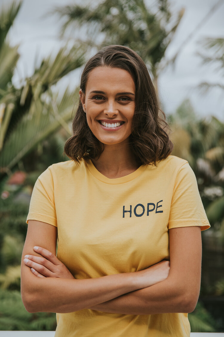 Image shows Genevieve smiling at the camera. She is wearing a yellow shirt with the word: "hope" on the front. She has shoulder-length brown hair and has her arms crossed in front of her.