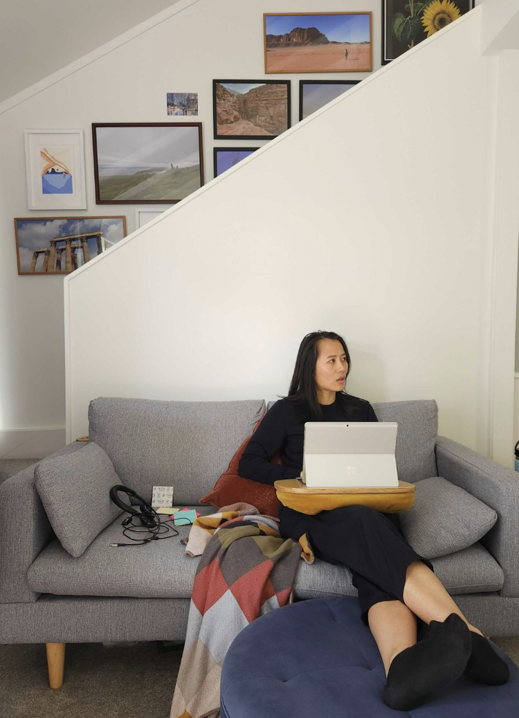 Image shows Yvonne sitting in her living room on a grey couch with a multicoloured patchwork blanket, her laptop on her lap and behind her are different pieces of art hanging up on the wall. She is wearing a black top, black pants and has long black hair.