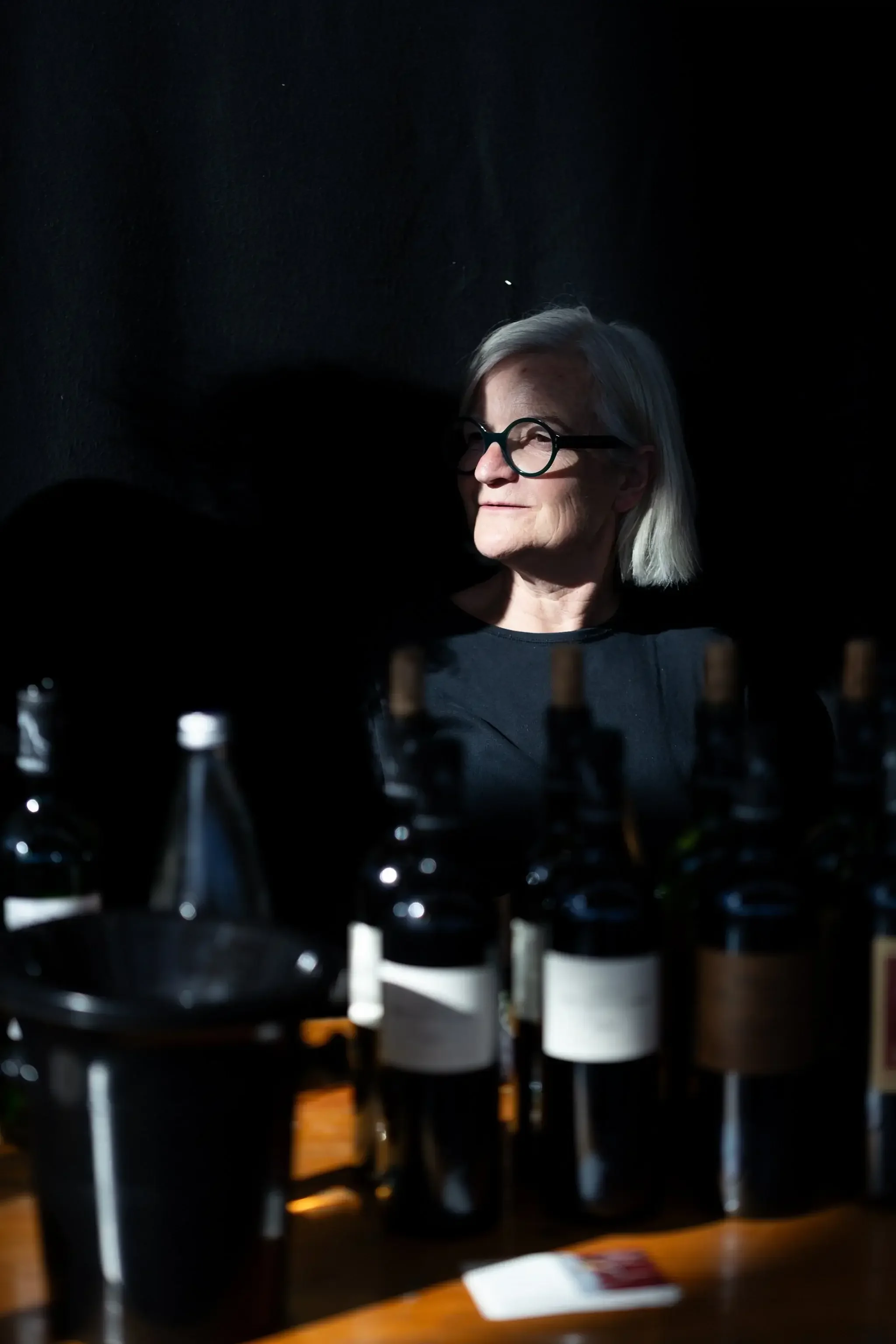 Une femme âgée avec des cheveux blancs, portant des lunettes noires, se tient à côté de plusieurs bouteilles de vin sur une table, dans un endroit sombre avec une lumière concentrée sur son visage.