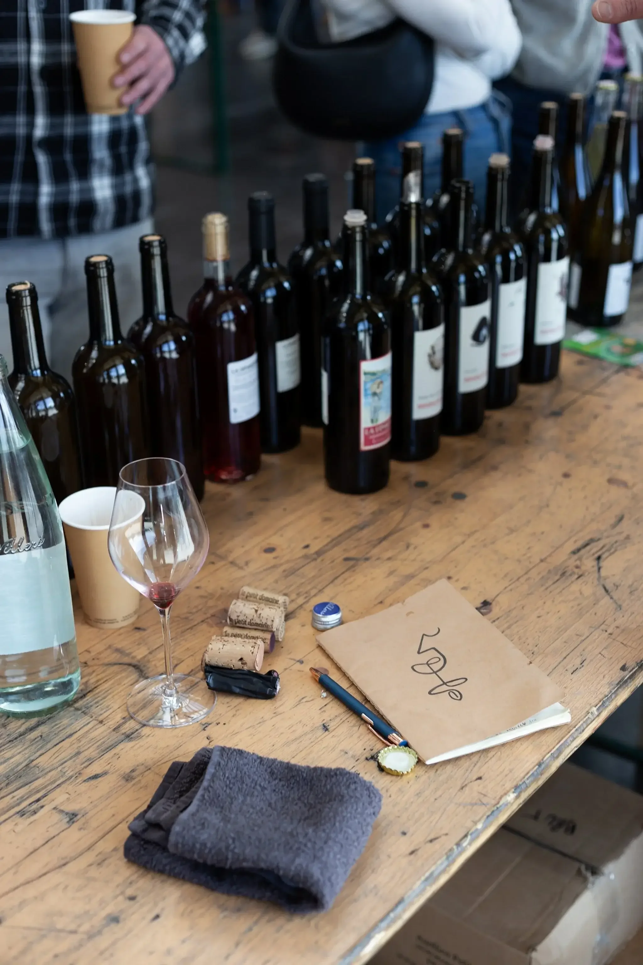 Différentes bouteilles de vin alignées sur une table en bois, avec un verre à vin vide, un carnet, une clé, des bouchons de vin, un marque-page, et une serviette en tissu. Des personnes sont visibles en arrière-plan.