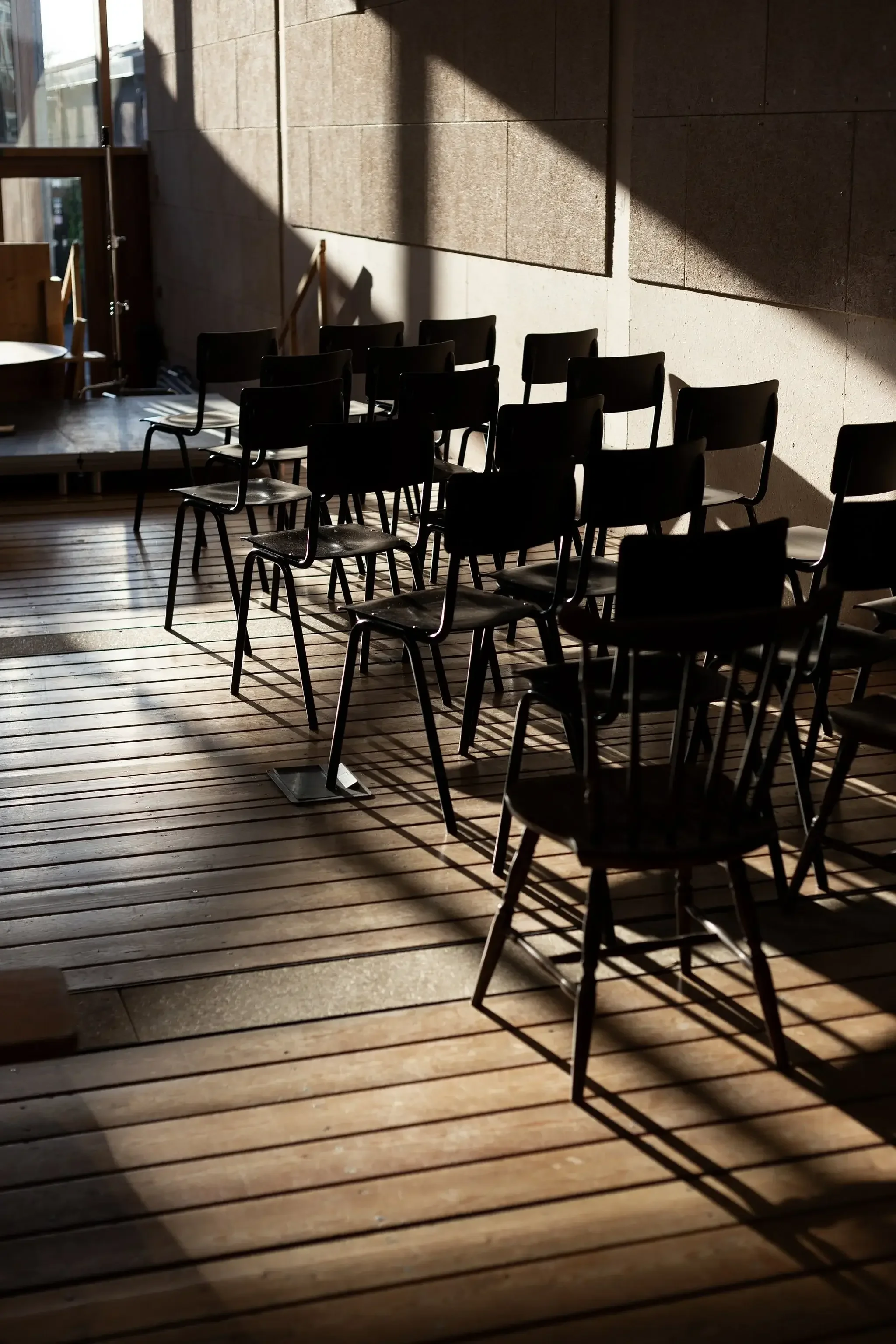 Chaises noires empilées dans une salle éclairée par la lumière naturelle, avec des ombres longues sur un sol en bois.