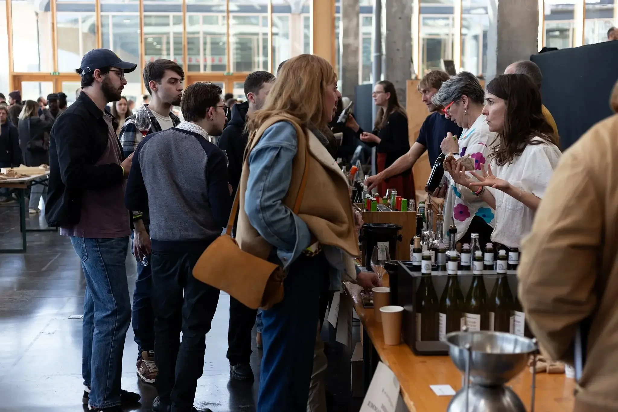 Gros plan d'une file de personnes faisant la queue pour déguster ou acheter des boissons lors d'un événement. Une jeune femme avec un sac marron, une veste en jean et une chemise à carreaux, écoute attentivement une autre personne derrière le stand o
