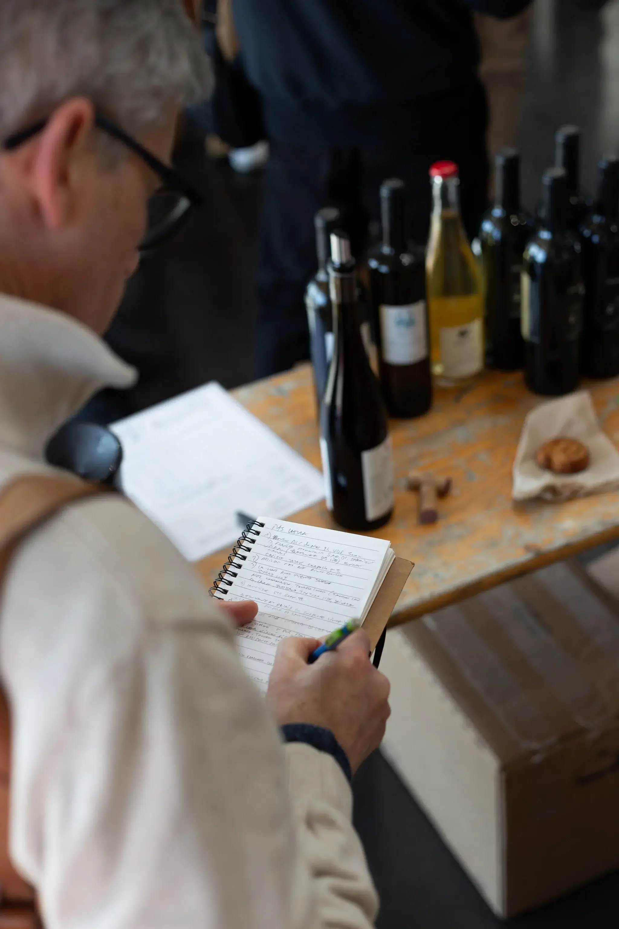 Un homme qui prend des notes dans un cahier à côté d'une table avec plusieurs bouteilles de vin, un biscuit et des documents.